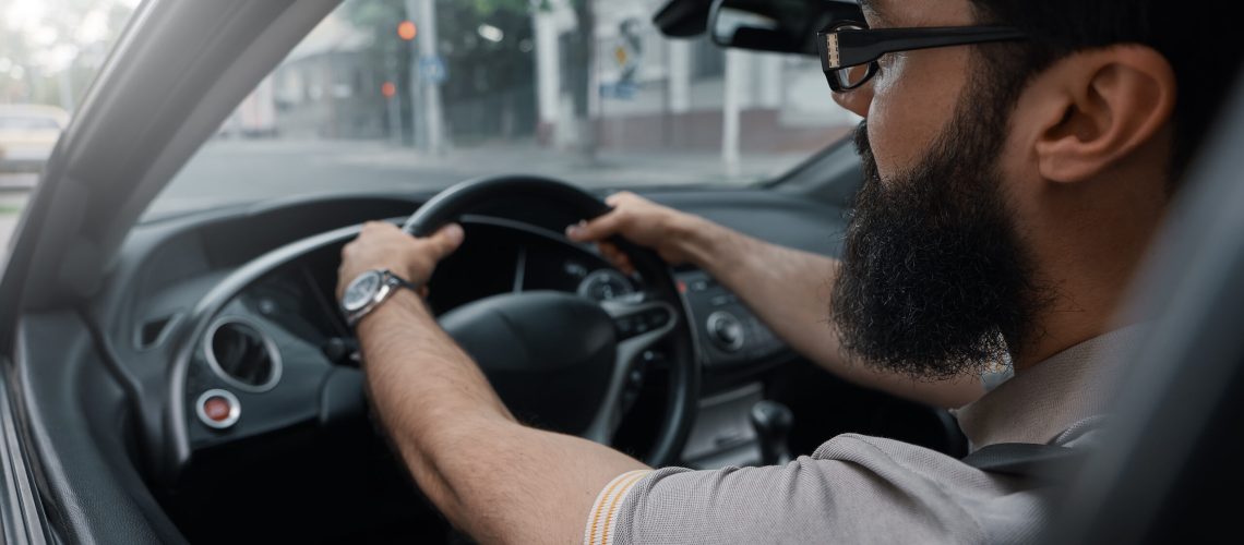 Angled side view modern casual bearded man, wearing glasses, watch and T-shirt, driving the car. Close up drivers' hands.